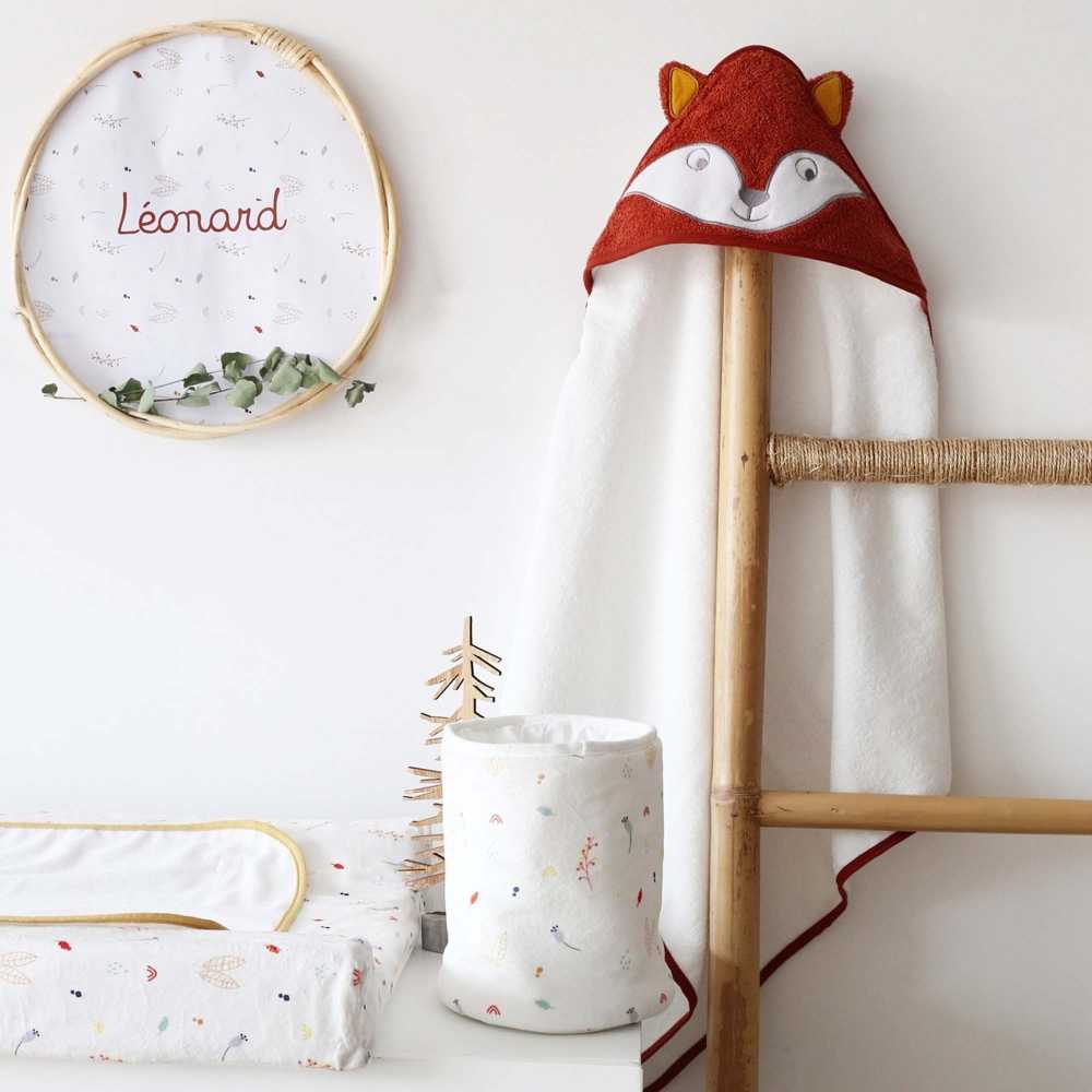 Pequeno saco com produtos para bebé pousado na cómoda, junto à capa de banho com raposa Pequeno saco com produtos para bebé pousado na cómoda, junto à capa de banho com raposa