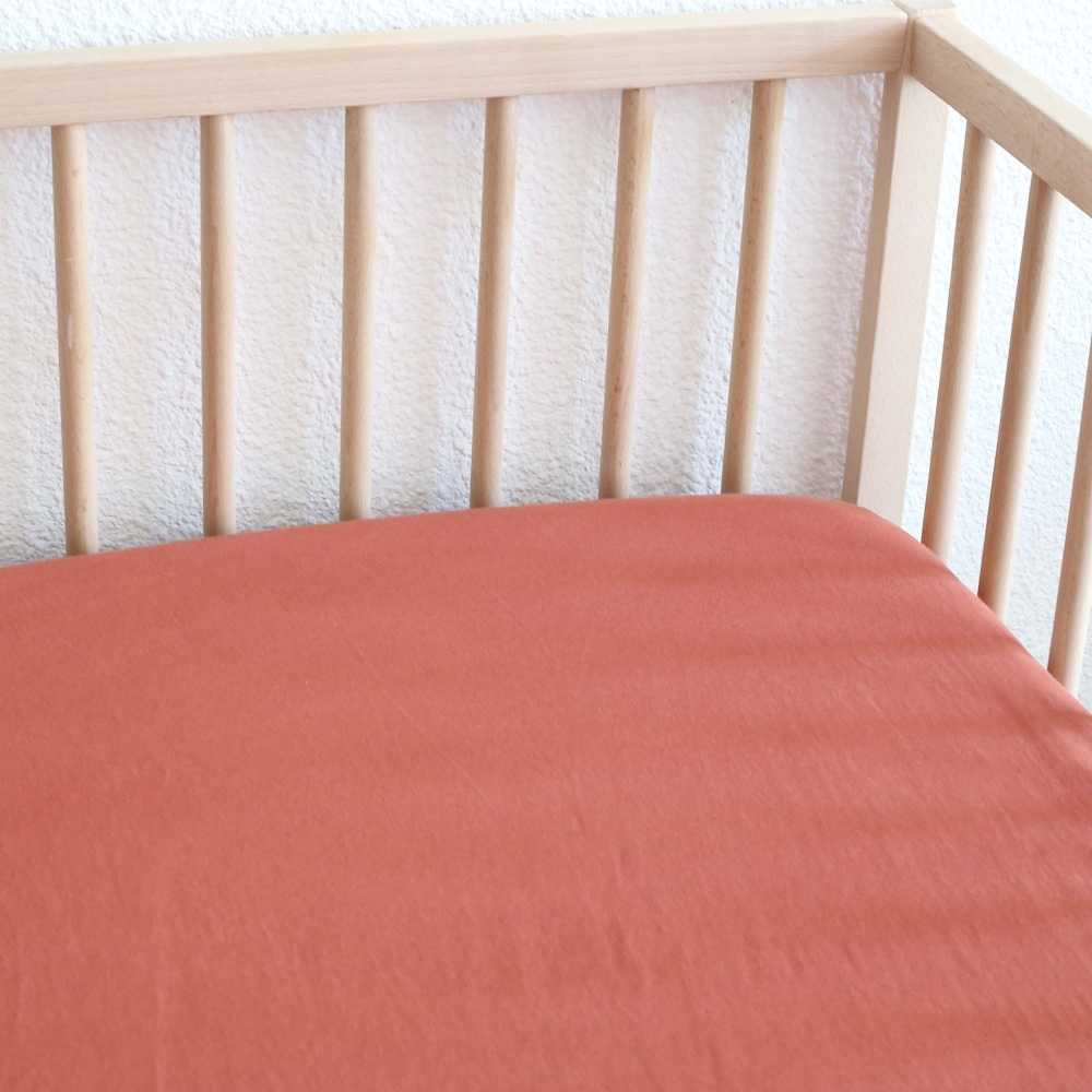 Lençol de baixo ajustável para bebé, liso, em tom terracota, numa cama de madeira Lençol de baixo ajustável para bebé, liso, em tom terracota, numa cama de madeira