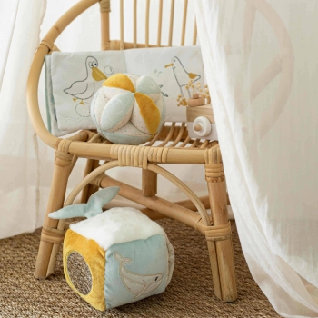 Cubo sensorial pousado no chão, com os outros brinquedos de estimulação da coleção numa cadeira de rotim Cubo sensorial pousado no chão, com os outros brinquedos de estimulação da coleção numa cadeira de rotim